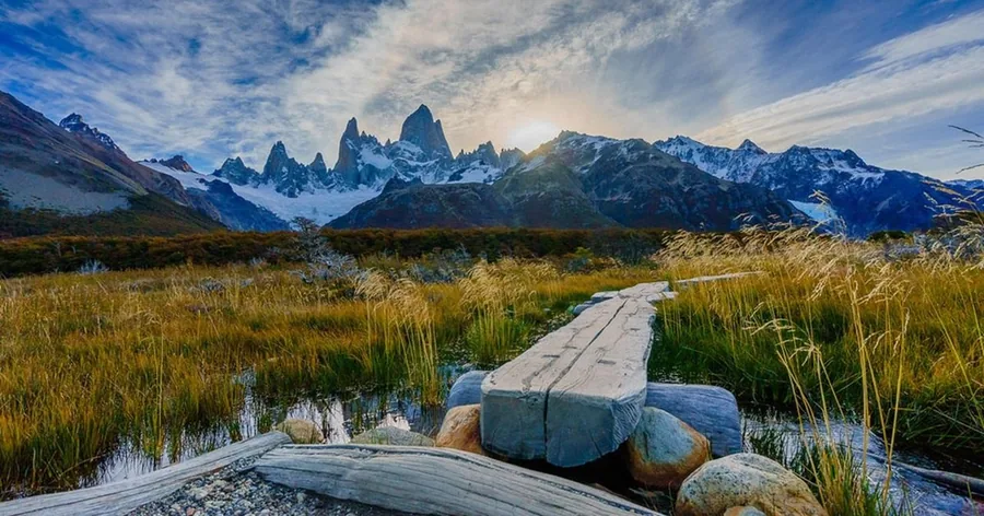 Ein Holzsteg über eine goldene Wiese in Richtung des verschneiten Fitz-Roy-Massivs an einem klaren Tag
