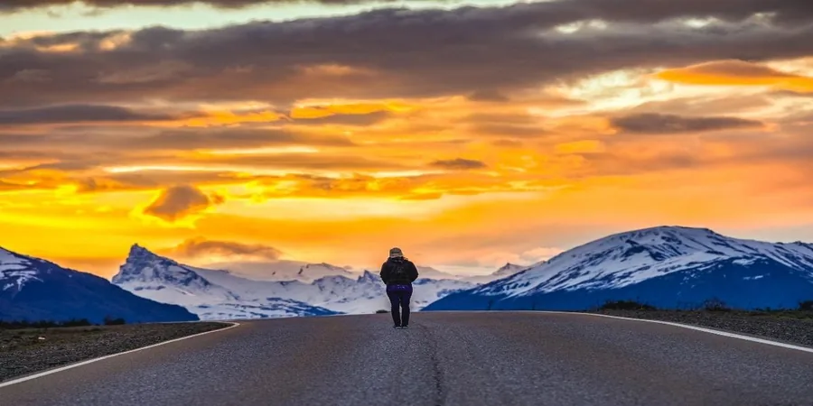 Ein einsamer Wanderer auf einer geraden Straße bei Sonnenuntergang, verschneite Gipfel am Horizont