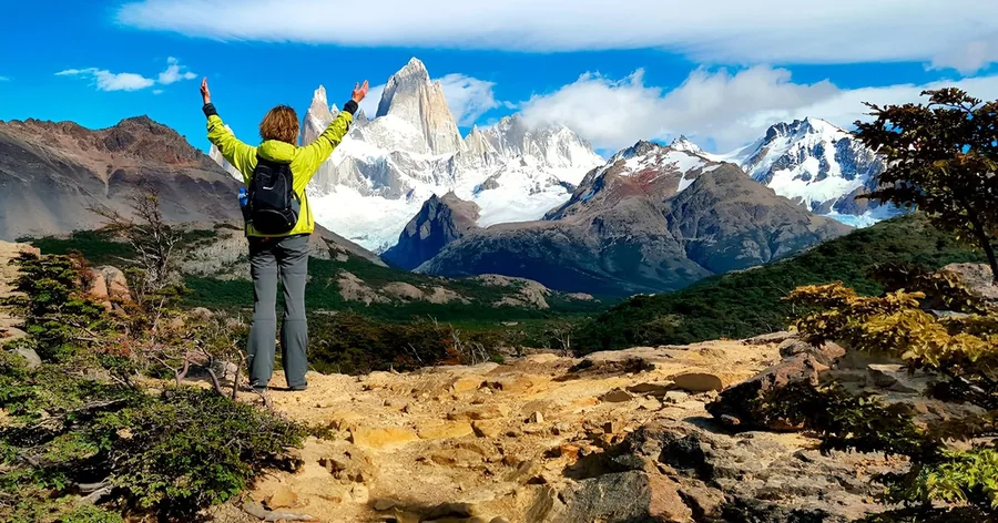 Ein Wanderer mit erhobenen Armen an einem felsigen Aussichtspunkt vor den Granitnadeln des Fitz Roy